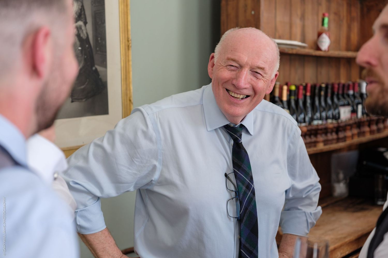 Elderly man in blue shirt and tie laughing joyfully during a casual conversation in a bar setting.