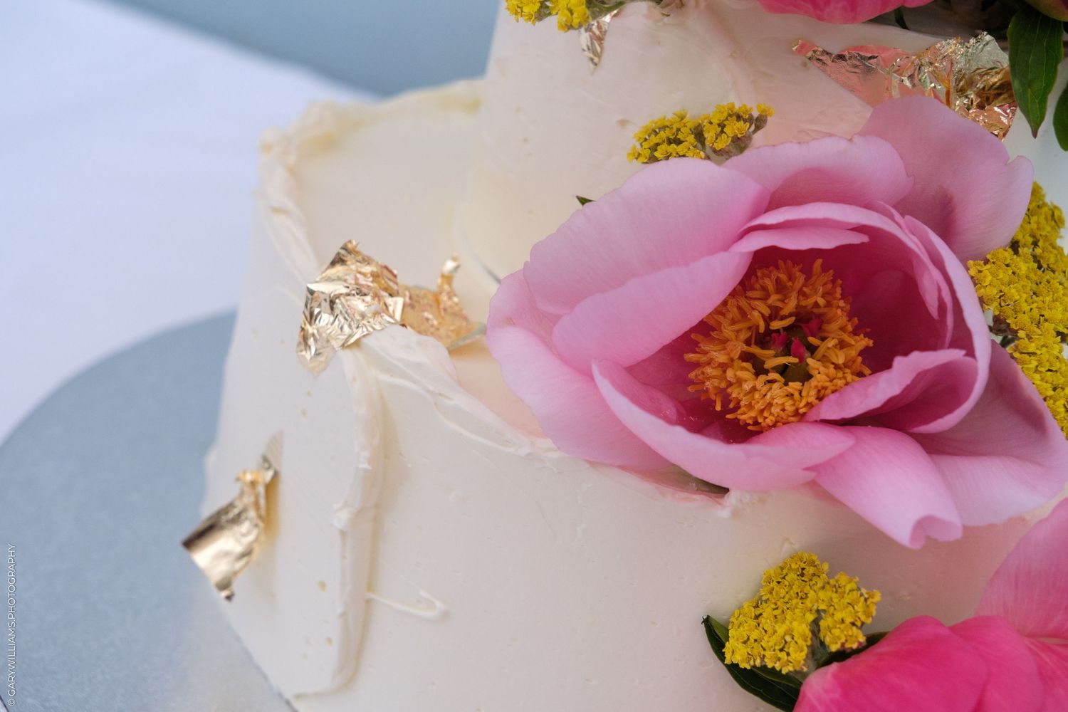 Close-up of white wedding cake with gold leaf details, pink peonies, and yellow floral accents.
