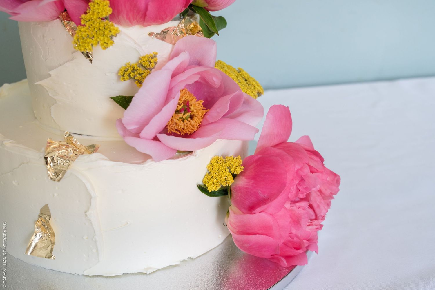 White tiered wedding cake decorated with pink peonies, yellow flowers, and gold leaf accents.