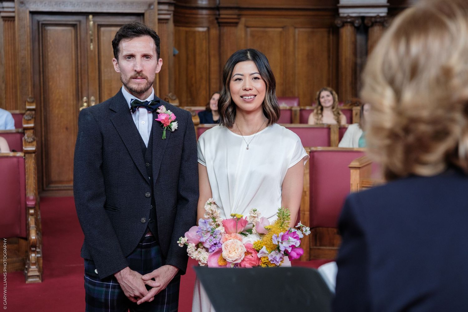 Couple stands at wedding altar, groom in tartan kilt with pink boutonniere, bride holding colorful bouquet.