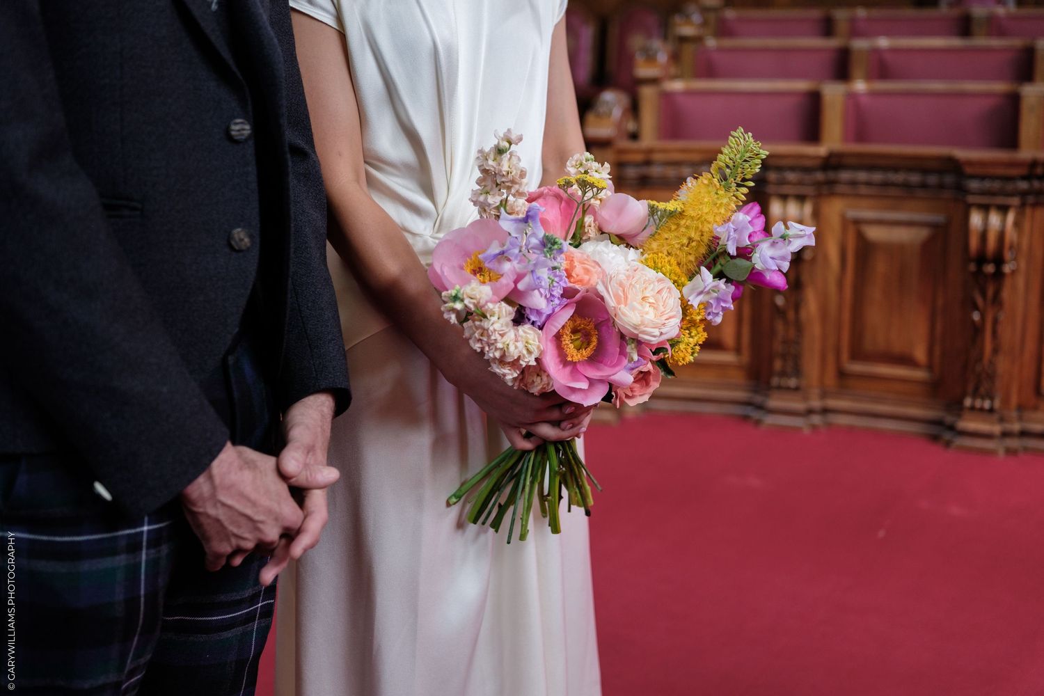 Bride holding a colorful spring wedding bouquet with pink, yellow, and purple flowers beside groom in tartan kilt.