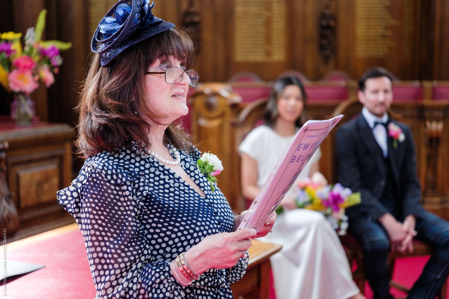Woman in polka dot dress reads from pink program at elegant wedding ceremony with couple seated behind her.