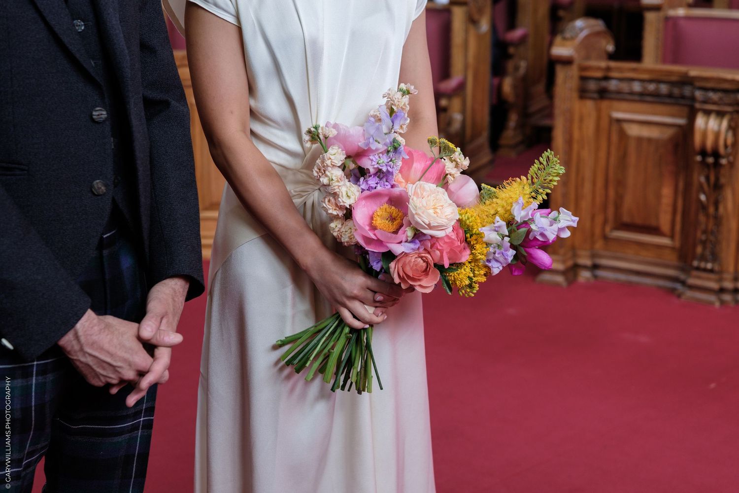 Bride holding colorful spring wedding bouquet with pink roses and yellow flowers, standing beside groom in tartan trousers.