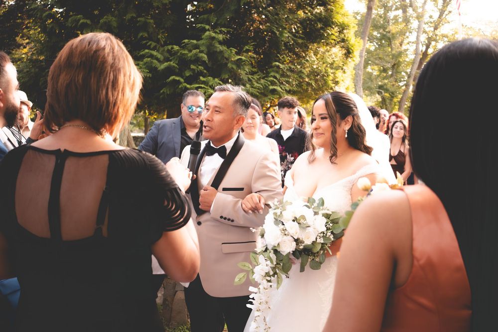 A bride holding a white floral bouquet walks with a man in a tan tuxedo at an outdoor wedding ceremony.