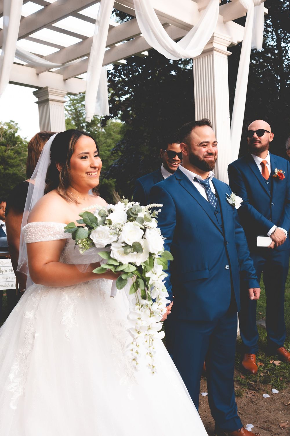 Bride in white off-shoulder gown holding floral bouquet stands beside groom in navy suit at outdoor wedding ceremony.