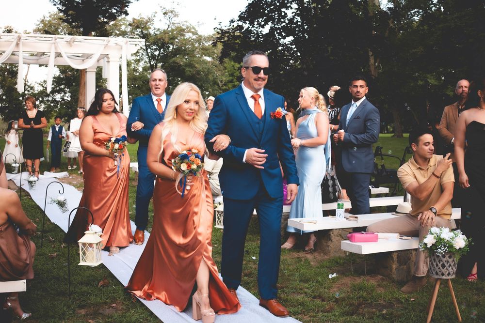 Bride in copper satin gown walks the aisle with man in blue suit at outdoor wedding ceremony with bridesmaids.
