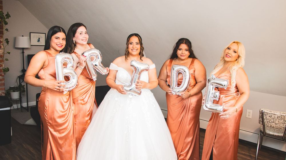Bride in white gown poses with four bridesmaids in rose gold dresses holding silver BRIDE balloon letters.