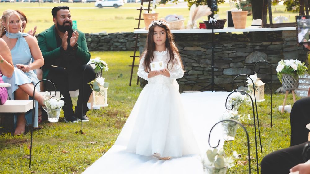 Young girl in white dress stands at outdoor wedding ceremony aisle with guests and officiant seated nearby.