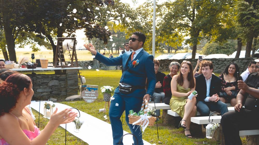 Man in blue suit speaking at outdoor wedding ceremony on green lawn.