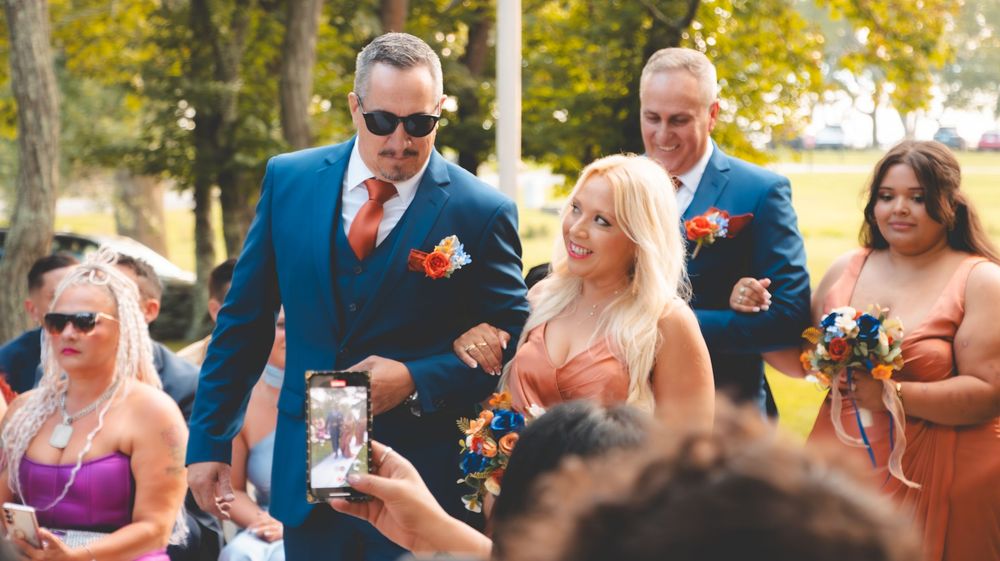 Groomsmen in blue suits with orange ties walk down the aisle at an outdoor wedding ceremony.