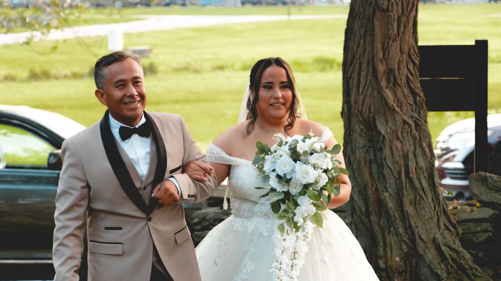 Bride in white gown holding bouquet walks with man in champagne tuxedo at outdoor wedding ceremony.