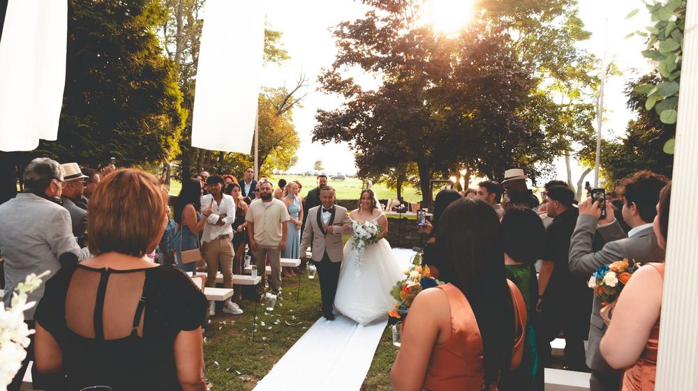Bride and groom walk down outdoor aisle surrounded by guests at a sunny garden wedding ceremony.