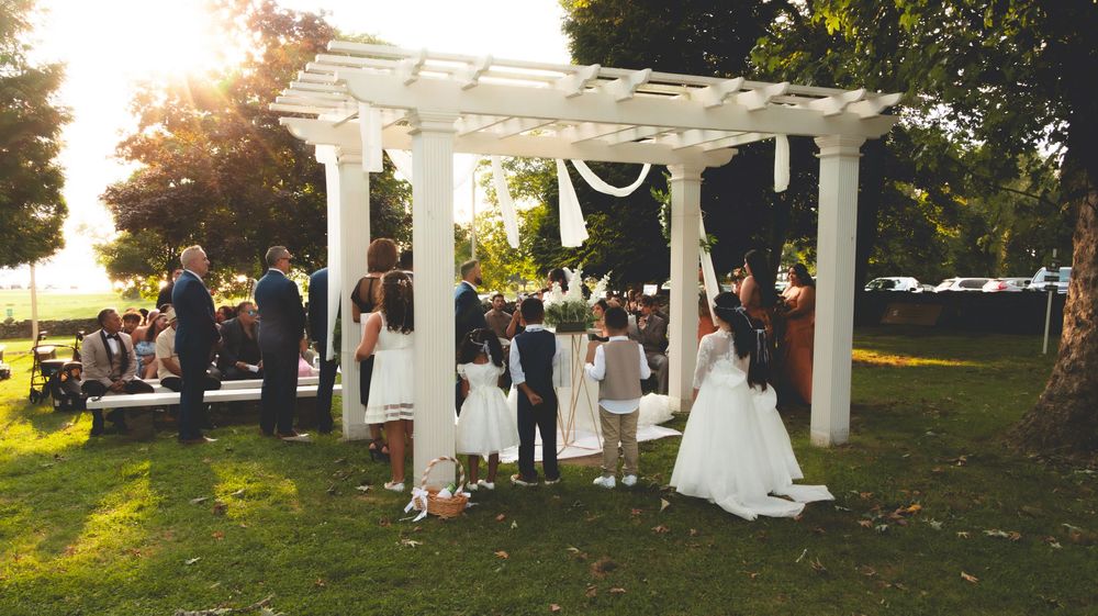 Wedding ceremony under white pergola at sunset with bridal party on lawn.