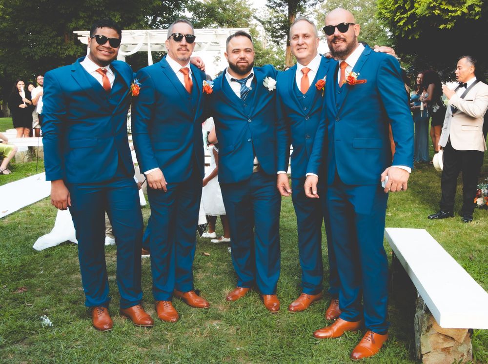Five groomsmen in matching blue suits with orange ties and brown dress shoes posing outdoors at a wedding reception.