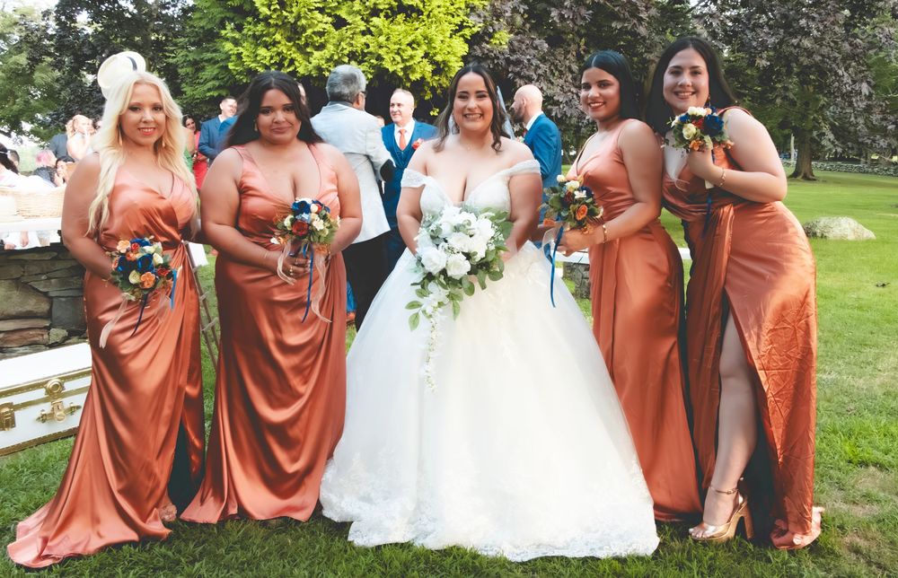 Bride in white gown poses with four bridesmaids in rust orange satin dresses holding bouquets at outdoor wedding ceremony.