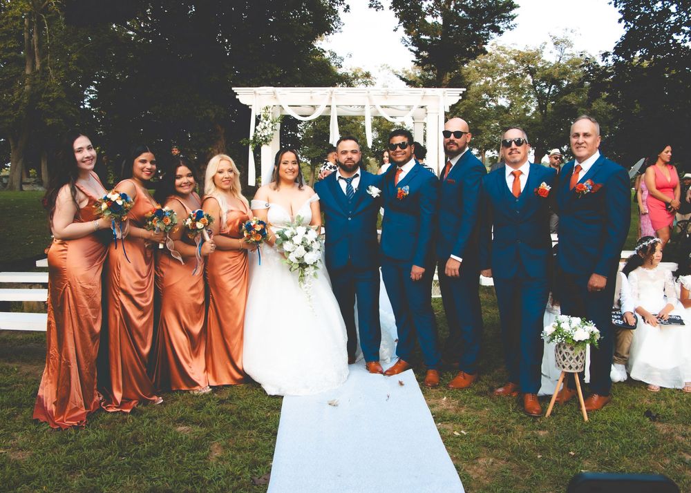 Wedding party posing outdoors with bridesmaids in orange dresses and groomsmen in navy suits beside bride and groom.