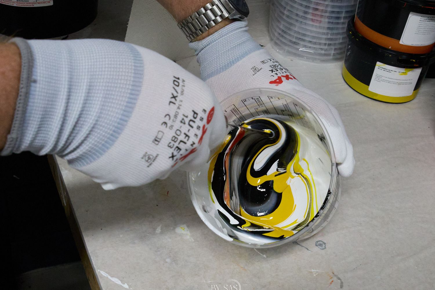 Gloved hands mixing black, white, and yellow paint swirls in a white bowl on a work surface.