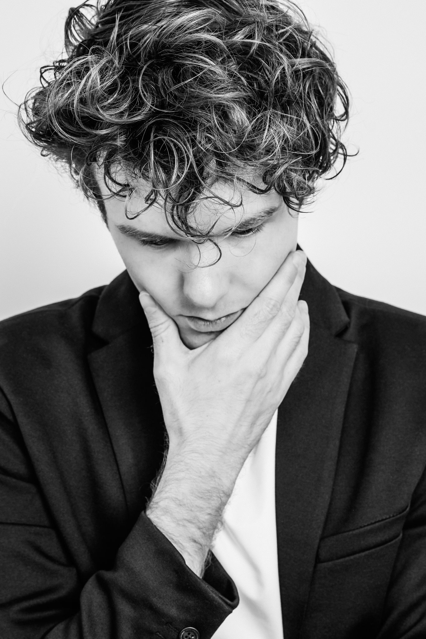Black and white portrait of a thoughtful young man with curly hair in a suit, hand on chin, looking down.