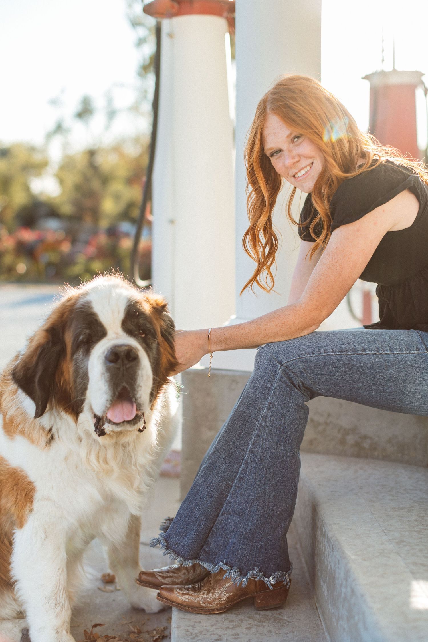 A red-headed young woman sits on concrete step in front of a vintage gas pump and leans forward, laughing as she pets her Saint Bernard.