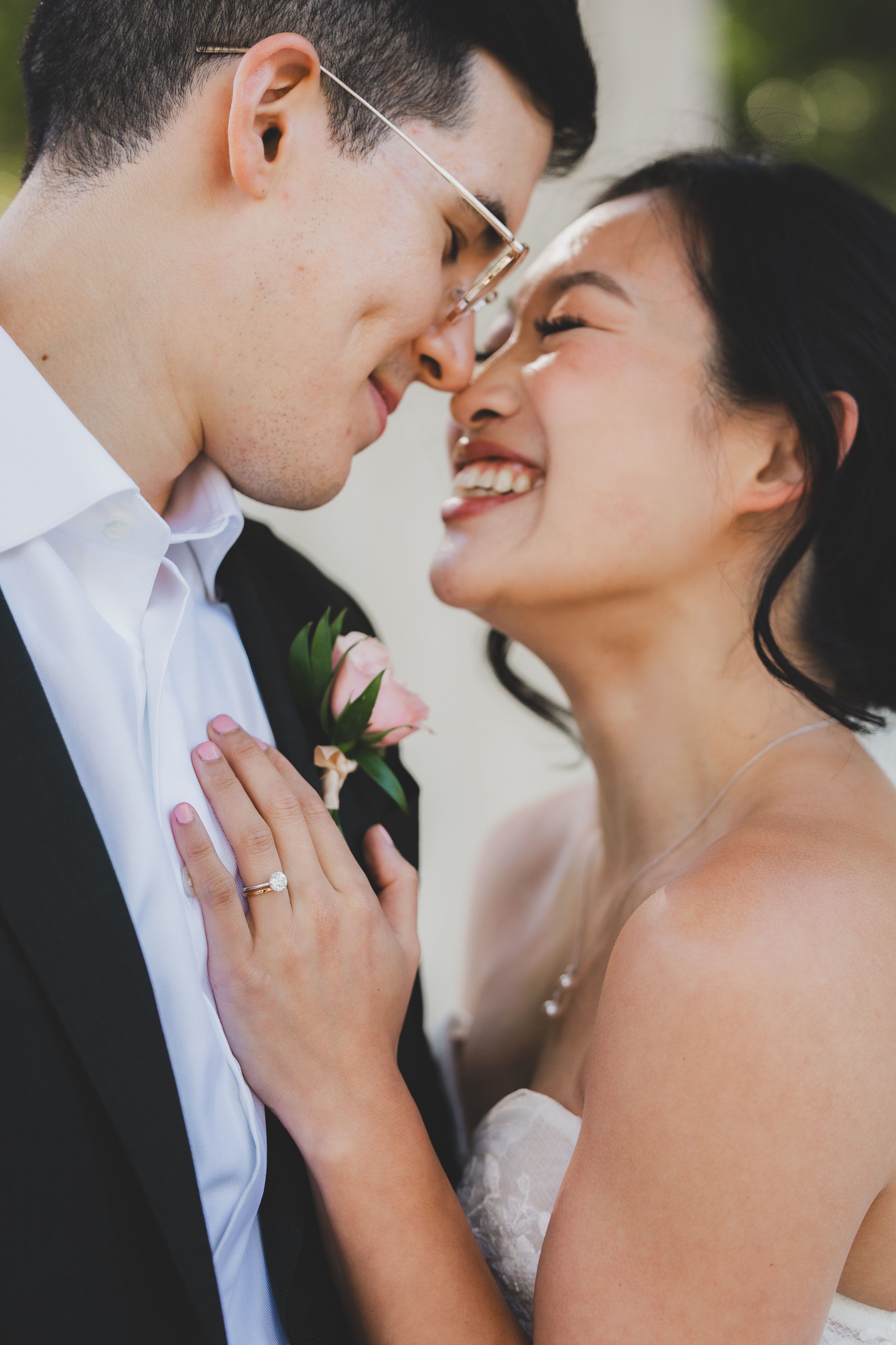 A joyful bride and groom share an intimate moment, touching noses and smiling, with a pink boutonniere and engagement ring visible.