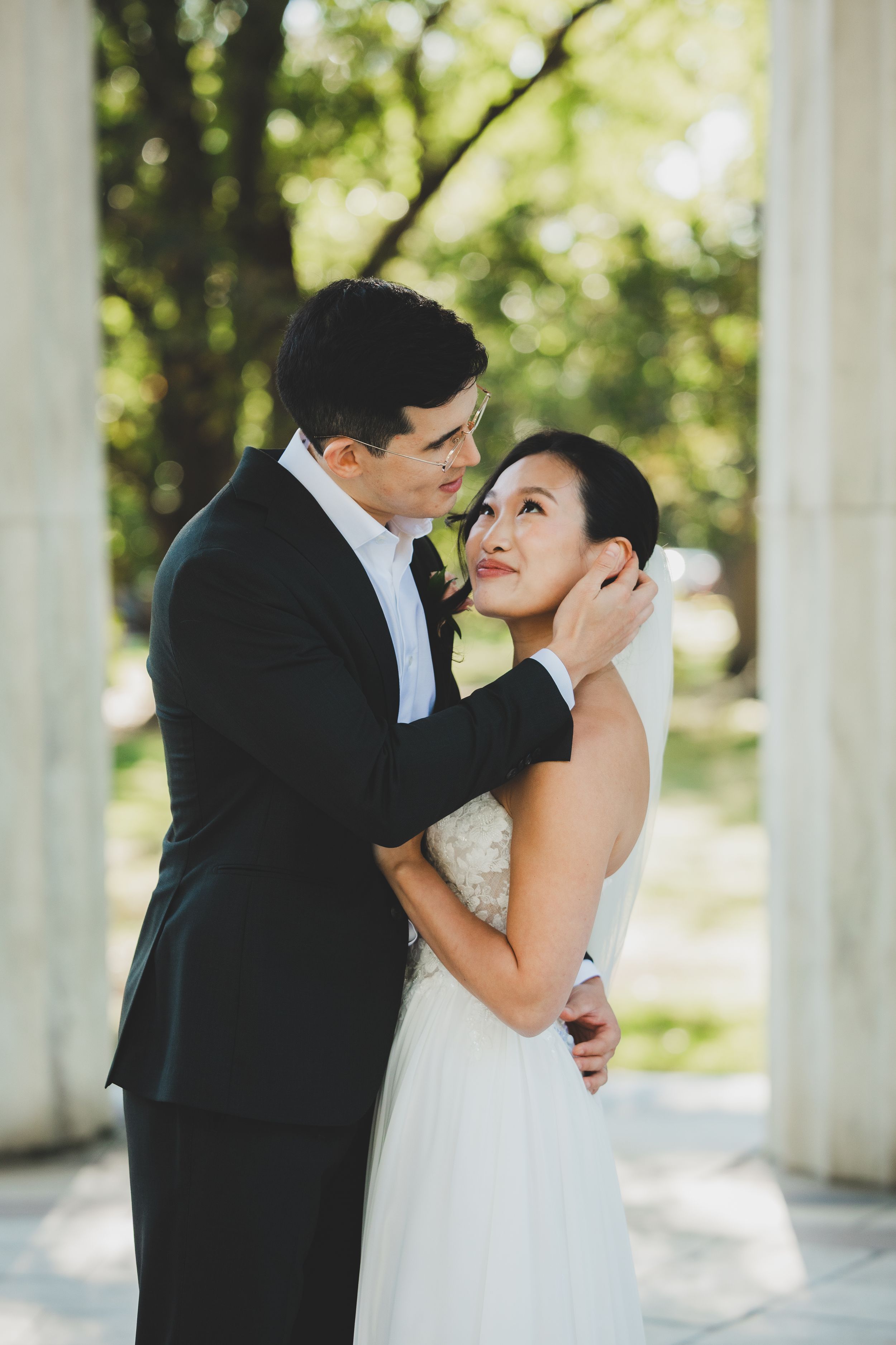 A groom in a black suit tenderly holds his bride in a white gown outdoors near white columns and lush green trees.