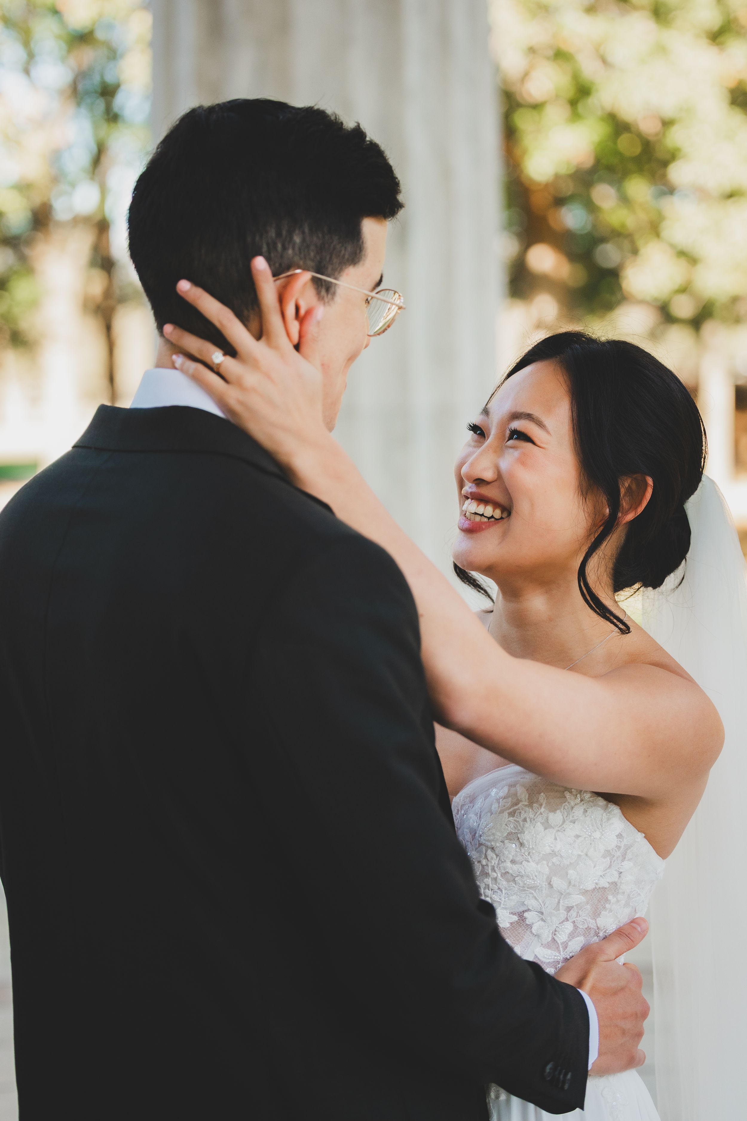 Smiling Asian bride in strapless lace gown embraces groom in black suit during outdoor wedding ceremony.