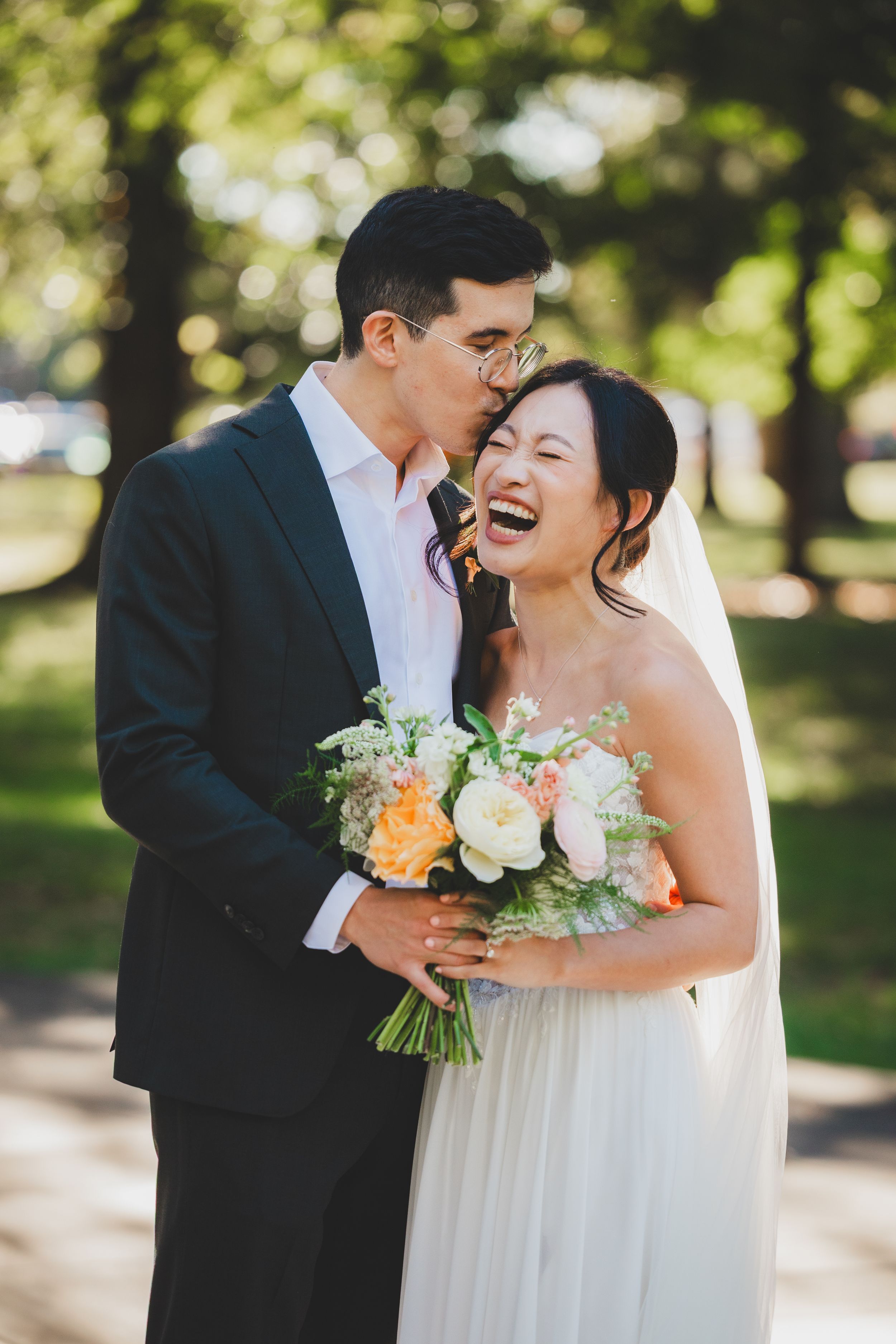 A joyful bride laughs as her groom kisses her cheek outdoors, she holds a peach and white floral bouquet.