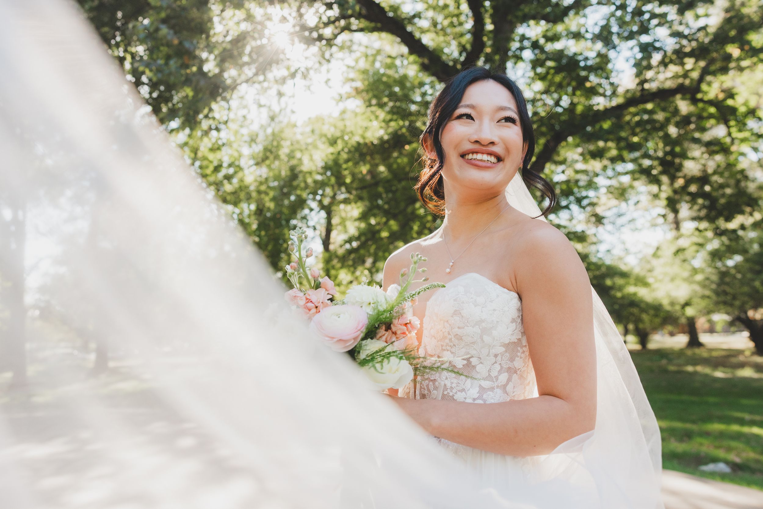 Smiling Asian bride in strapless lace gown holds pink bouquet outdoors, veil flowing in breeze with sunlit trees behind.