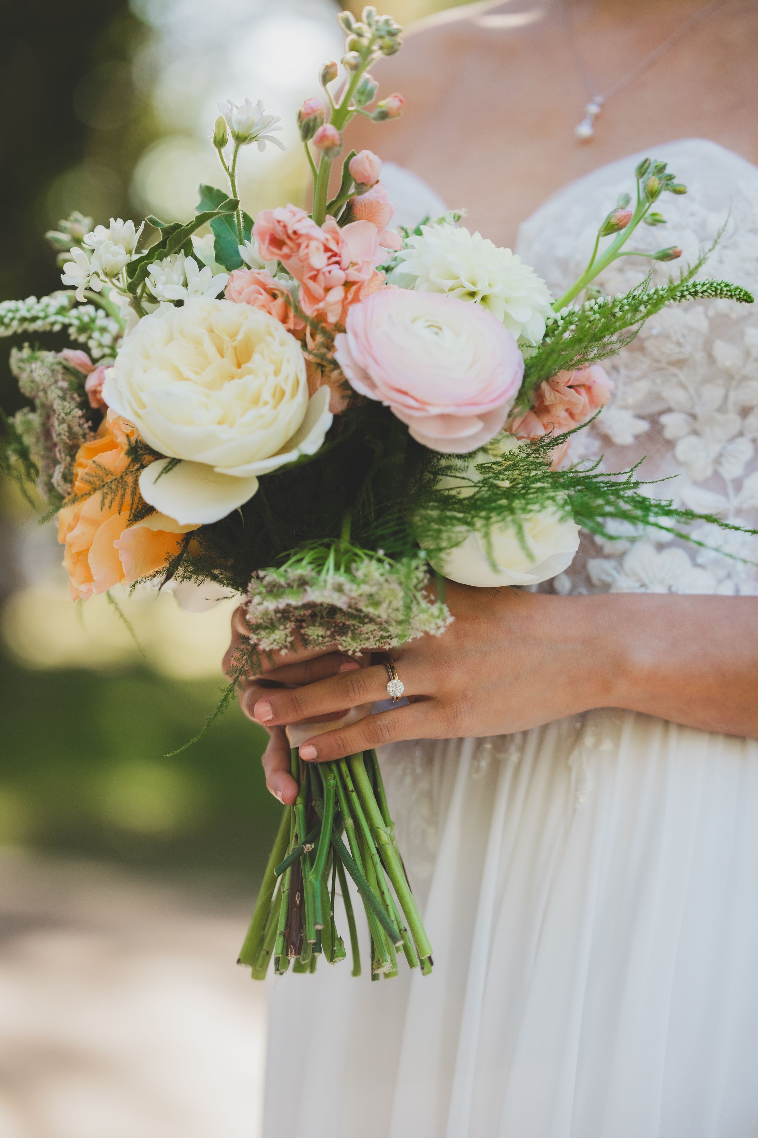 Bride holding a romantic wedding bouquet with peach, cream, and blush roses, greenery, wrapped in burlap ribbon.