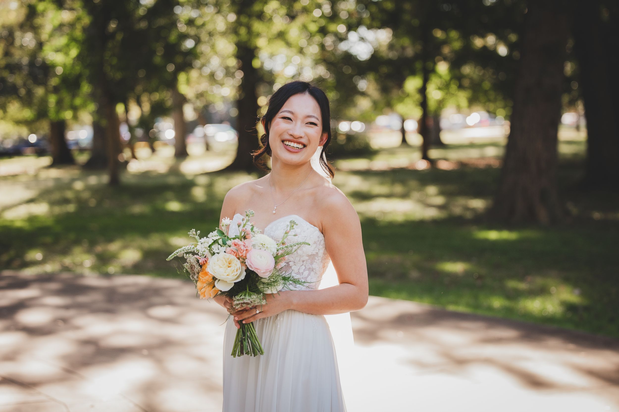 Smiling bride in strapless white gown holding pastel bouquet of flowers, standing in sunlit park with trees.