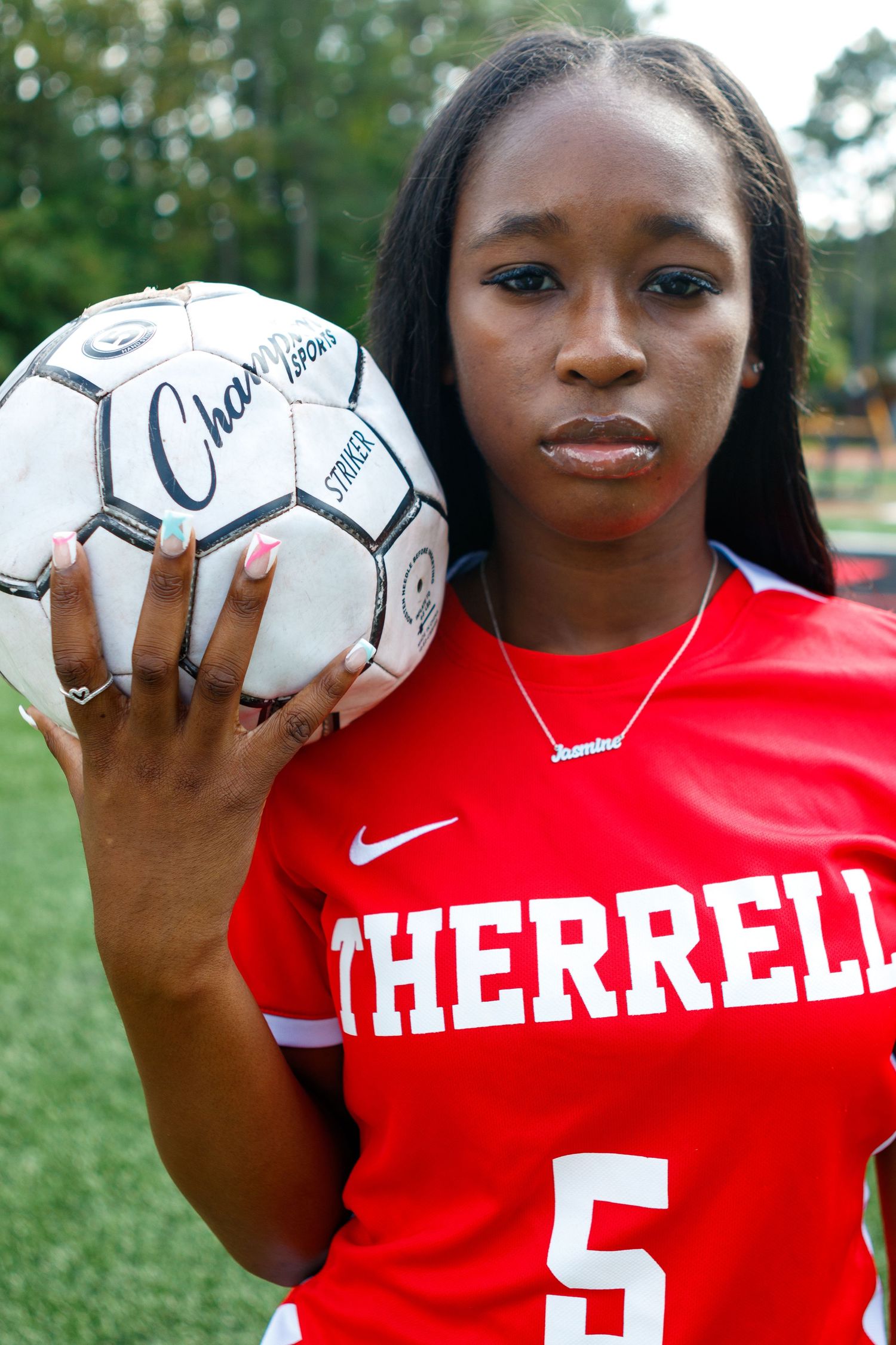Young female soccer player in red jersey number 5 holding a ball and pointing at camera.