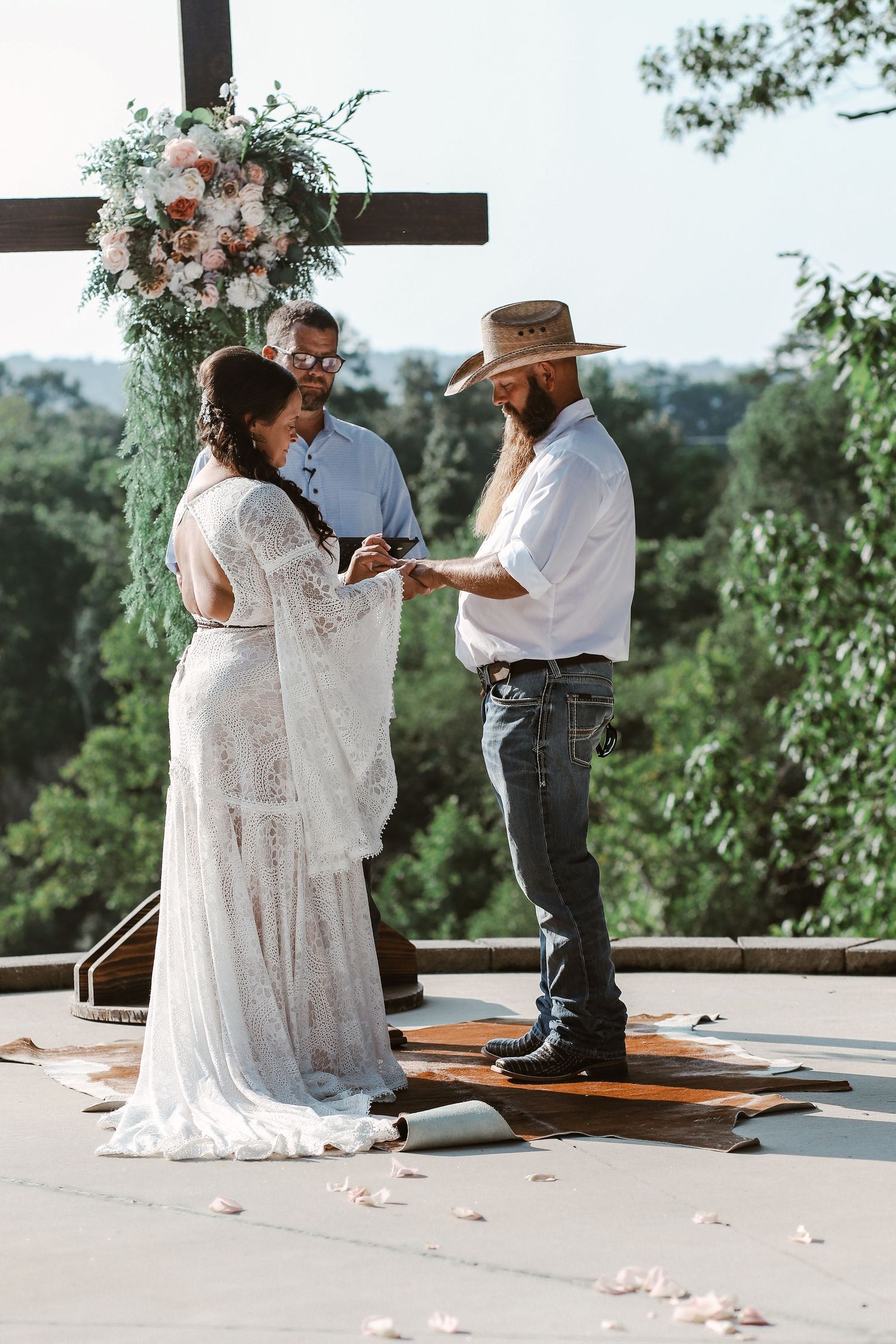 Bride and groom exchange vows outdoors at rustic boho wedding ceremony beneath floral cross with lush greenery backdrop.