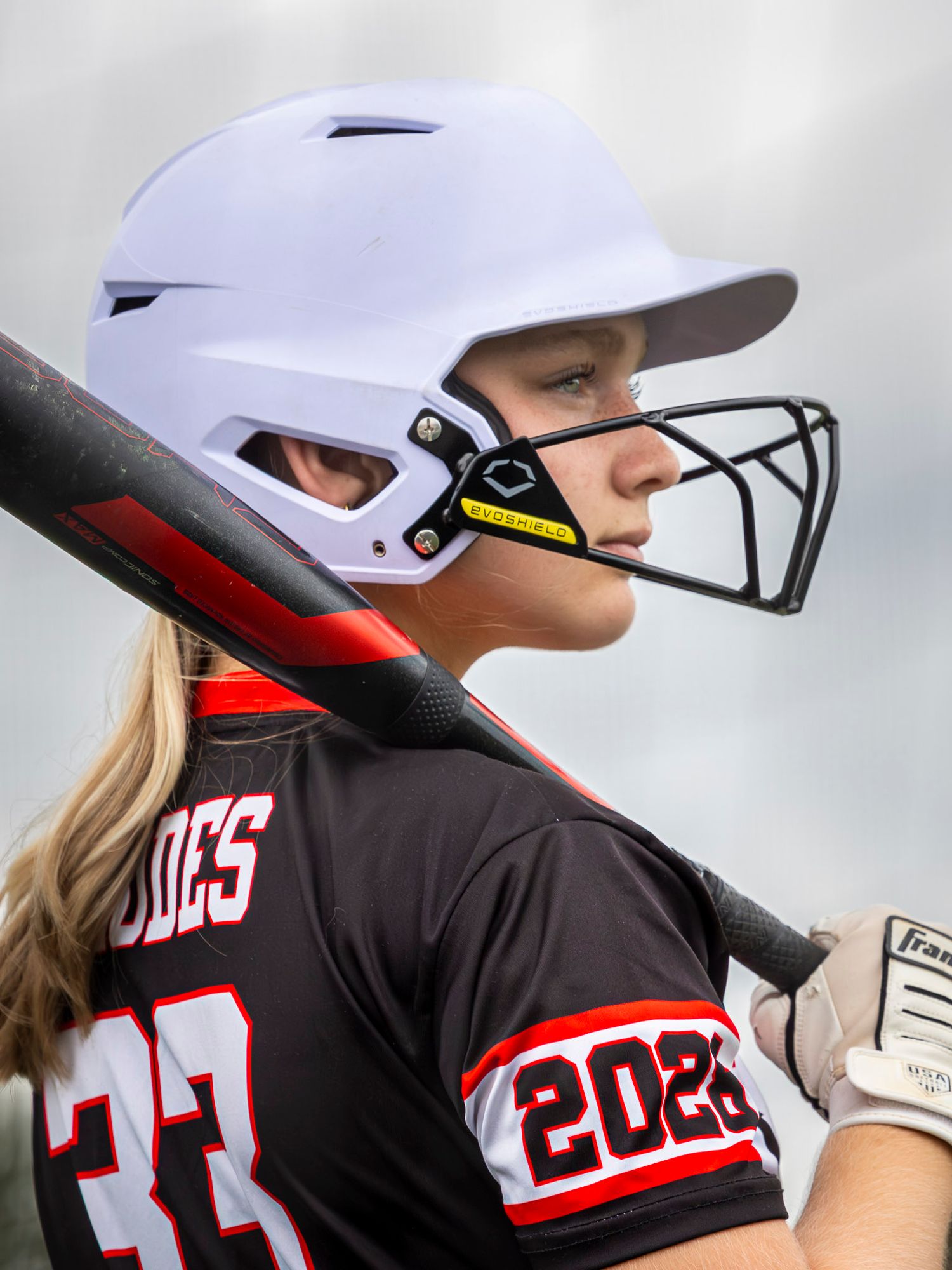 Female softball player wearing #33 jersey, white batting helmet with face guard, holding bat ready to hit.