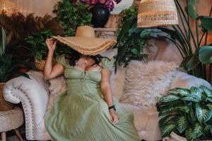 Woman in green dress and wide-brim hat relaxing on a couch surrounded by lush tropical plants indoors.
