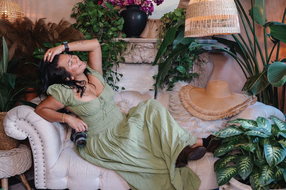 Woman in green dress lounging on white sofa surrounded by lush tropical plants indoors.