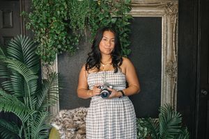 Woman in plaid dress holding camera surrounded by lush tropical plants and ornate mirror backdrop.