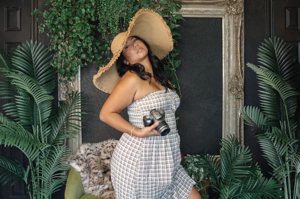 Woman in wide-brim hat and plaid dress holding camera, posing among tropical plants near decorative frame.