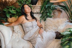 Woman in plaid jumpsuit relaxing on cream sofa surrounded by lush tropical plants in boho-style setting.