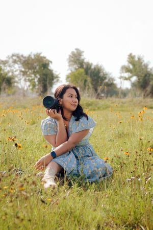 Female photographer in blue floral dress holding DSLR camera while sitting in wildflower field.