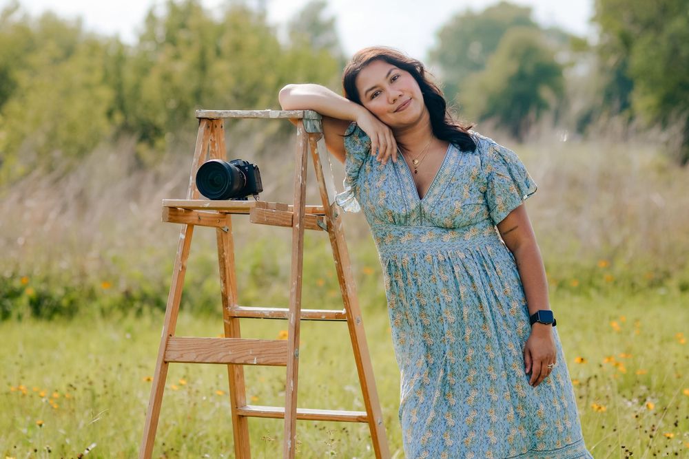 Woman in floral blue dress leaning on wooden ladder in field with camera, outdoor portrait photography.