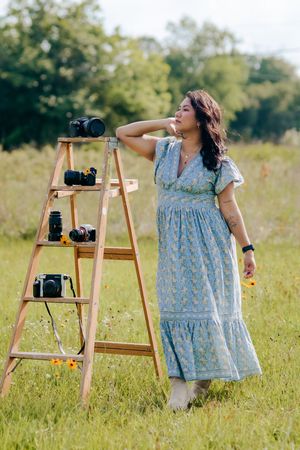 Female photographer in blue maxi dress standing beside camera-filled wooden ladder in green field.