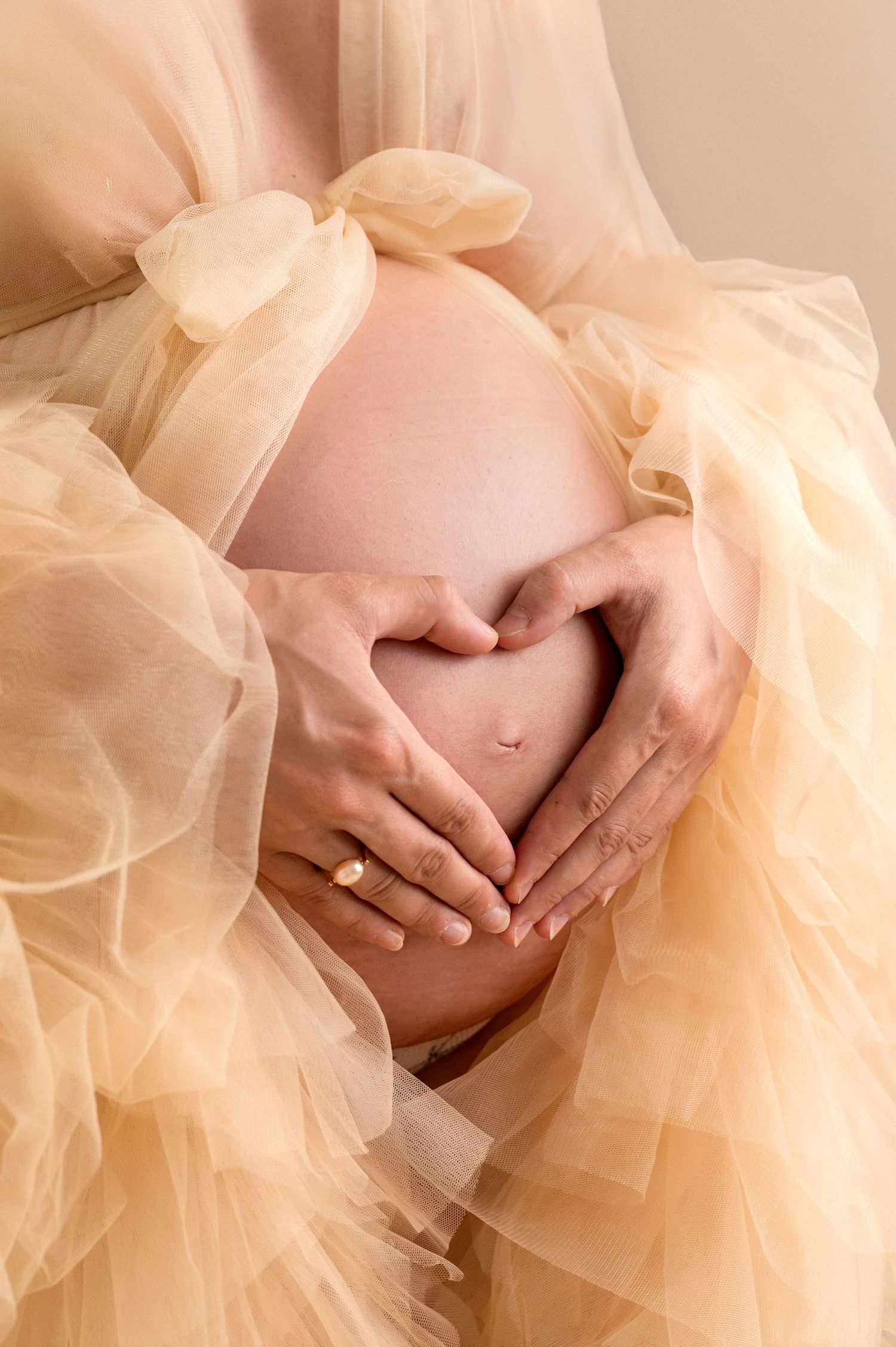 Pregnant woman in white lace maternity gown posing against floral gray backdrop in professional studio photo.