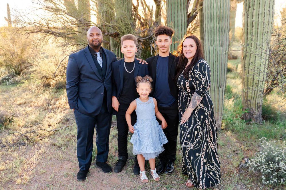 Family of five dressed in formal attire poses together among tall saguaro cacti at golden hour.