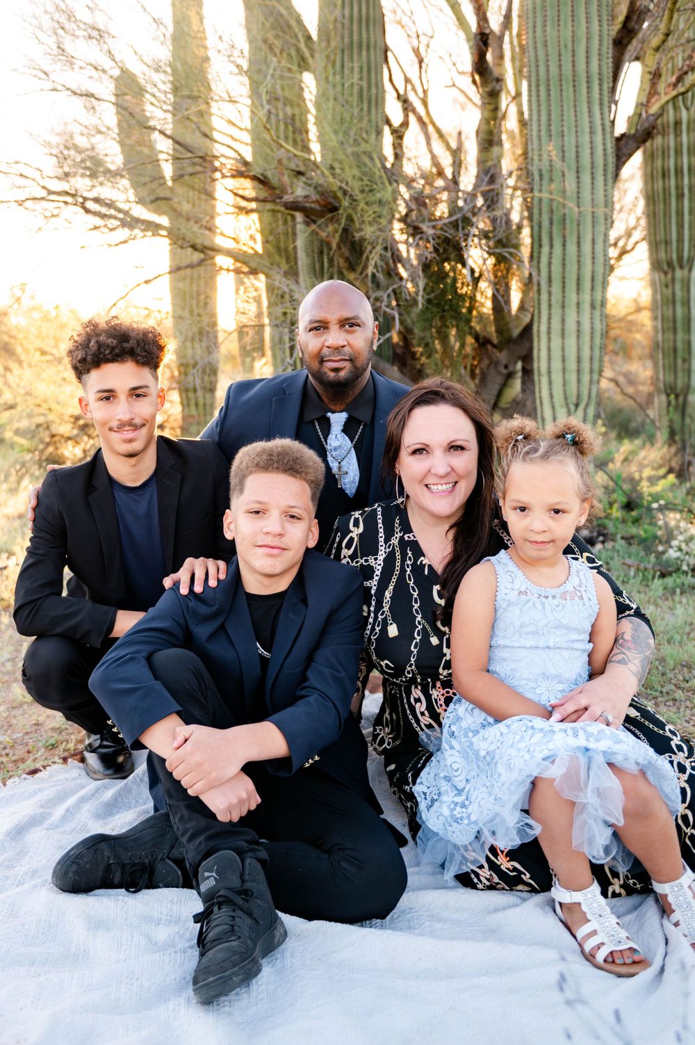 Family of five in coordinated dark outfits poses together near saguaro cactus at desert sunset.
