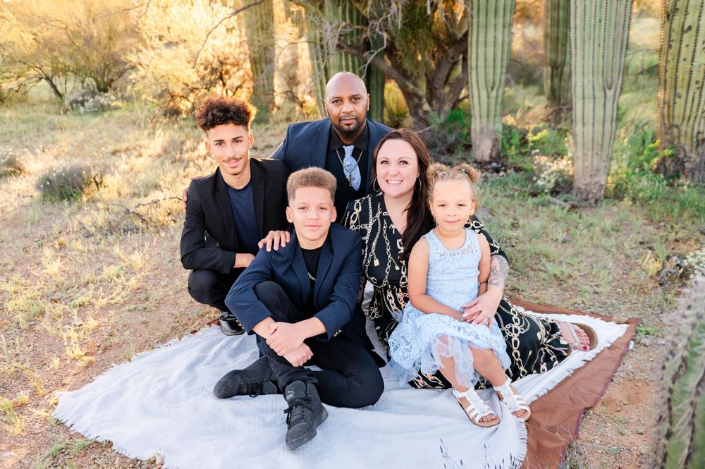 Family of five posing on white blanket in desert with saguaro cacti during golden hour photography session.