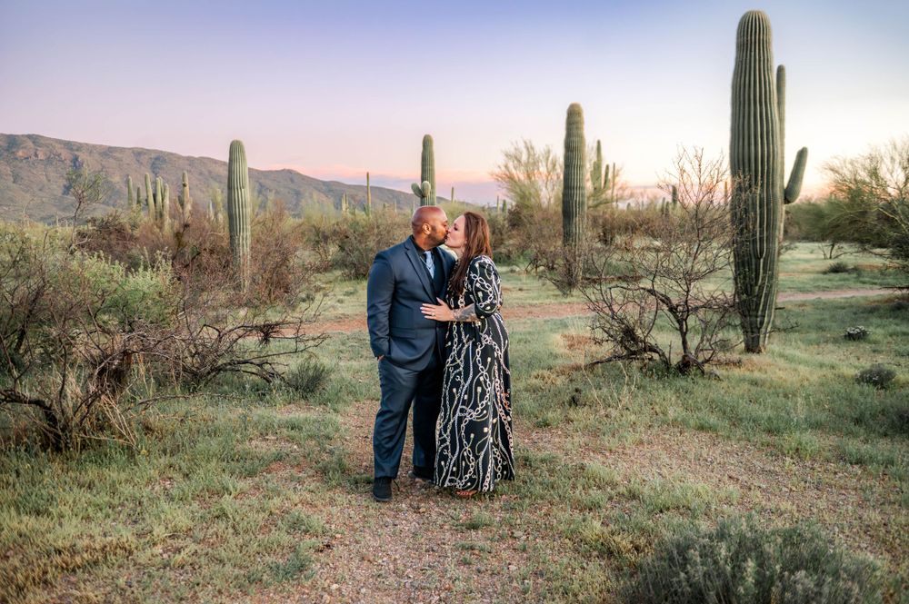 Couple sharing a romantic kiss surrounded by tall saguaro cacti in Arizona desert at sunset.