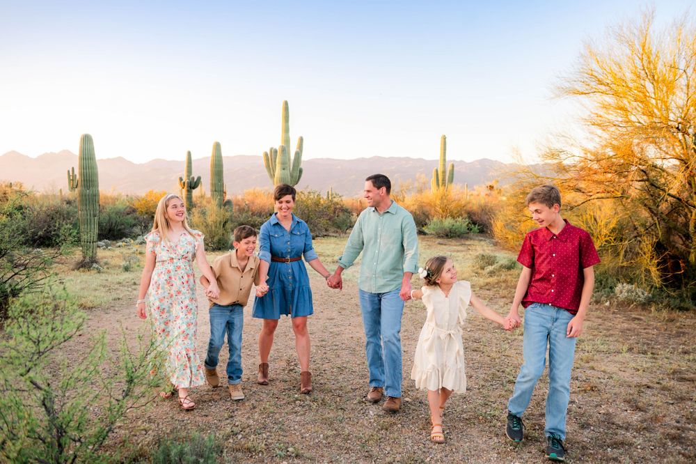 Family of six holding hands walking through desert landscape with saguaro cacti and golden sunset.