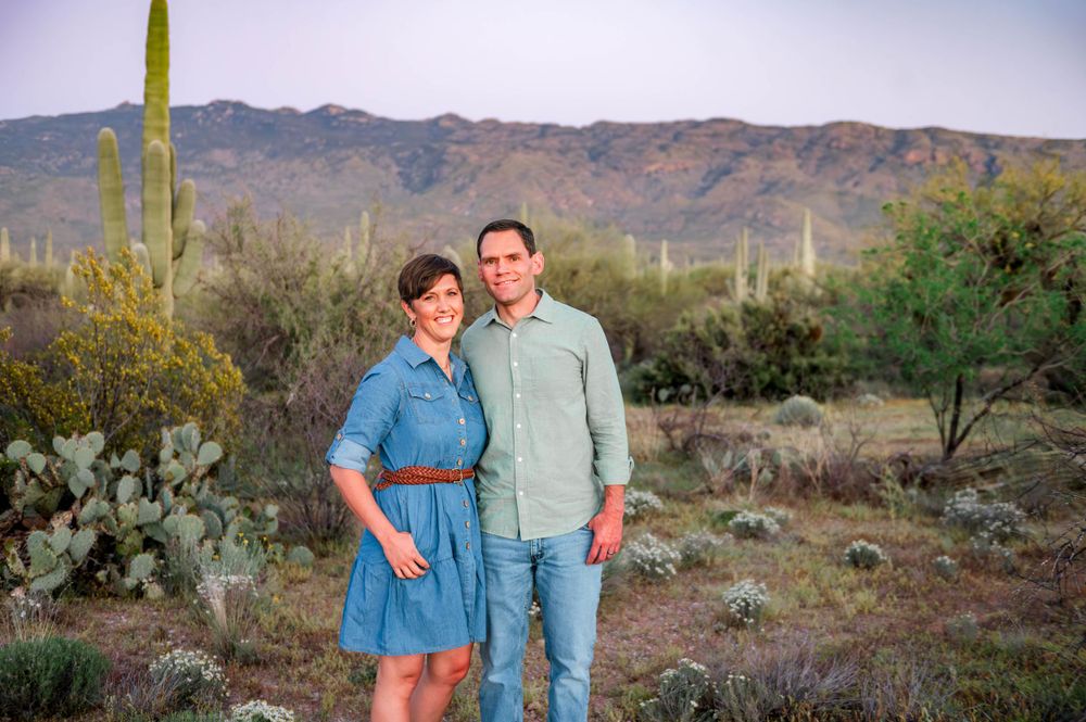 Smiling couple stands together in desert landscape with saguaro cacti and mountains at sunset.