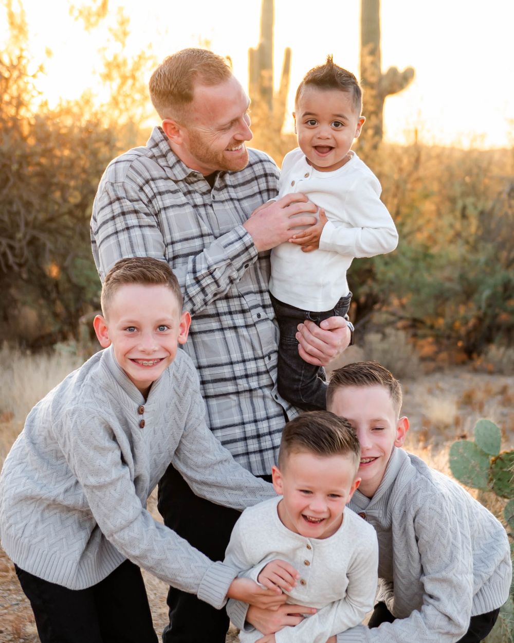 Father laughs with four young sons outdoors among saguaro cacti in warm golden sunset light.