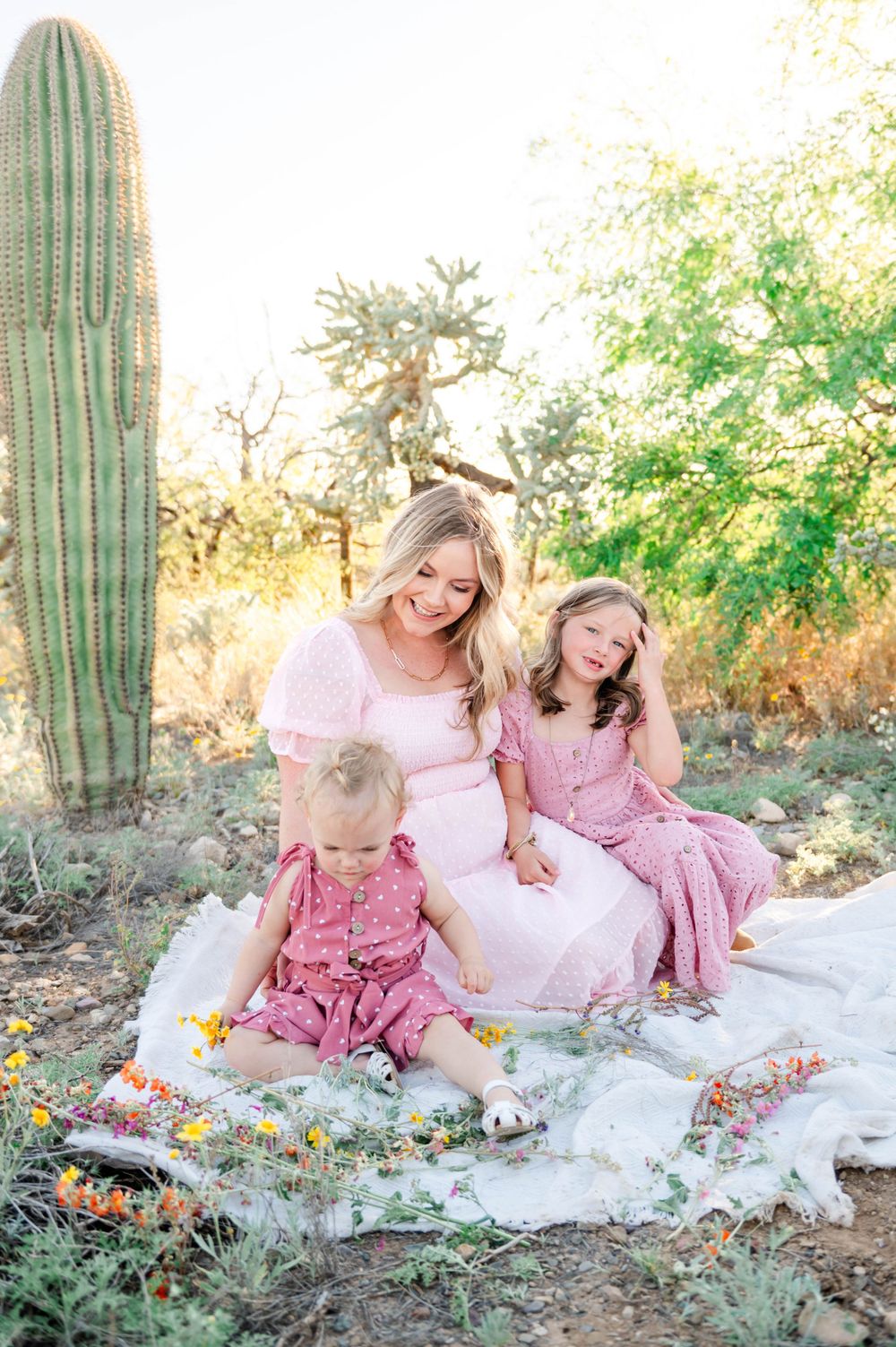 A pregnant woman in pink sits on a blanket with two young daughters near a saguaro cactus.